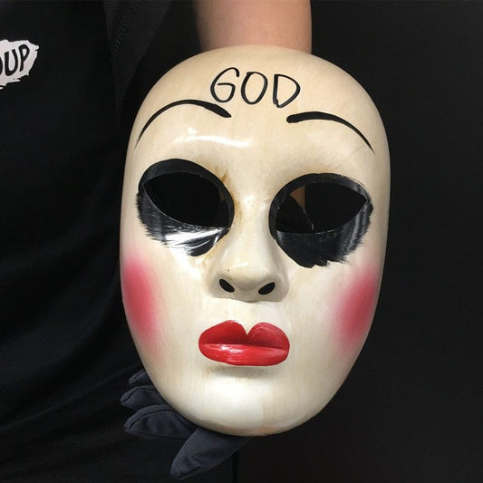 A hand holds a white mask with red lips, black eye holes, and "GOD" written on the forehead. The mask also features exaggerated eyebrows and smudged black makeup.