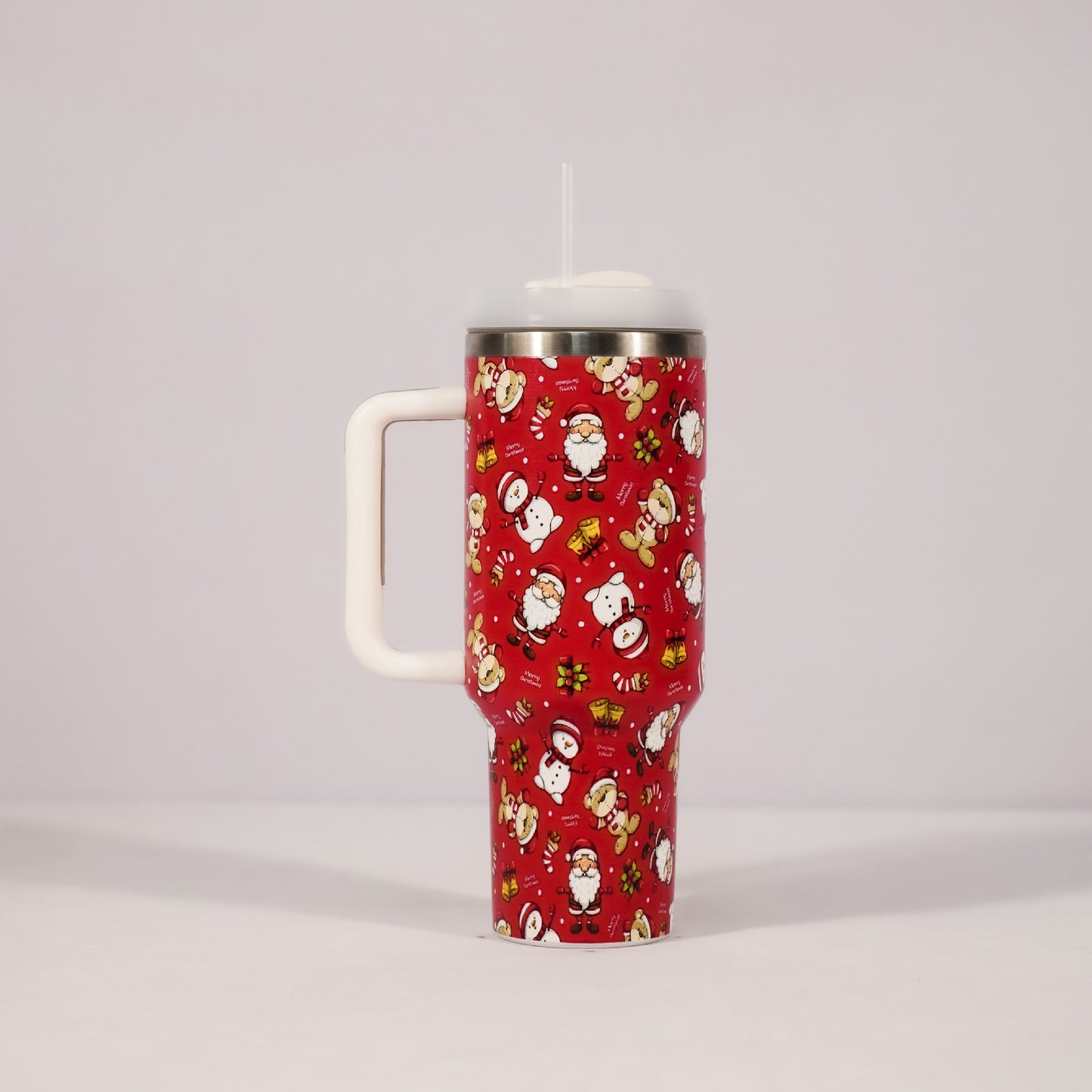 Red insulated tumbler with a straw, covered in festive holiday characters like Santa, snowmen, and reindeer, accompanied by candy canes and presents on a white handle backdrop.