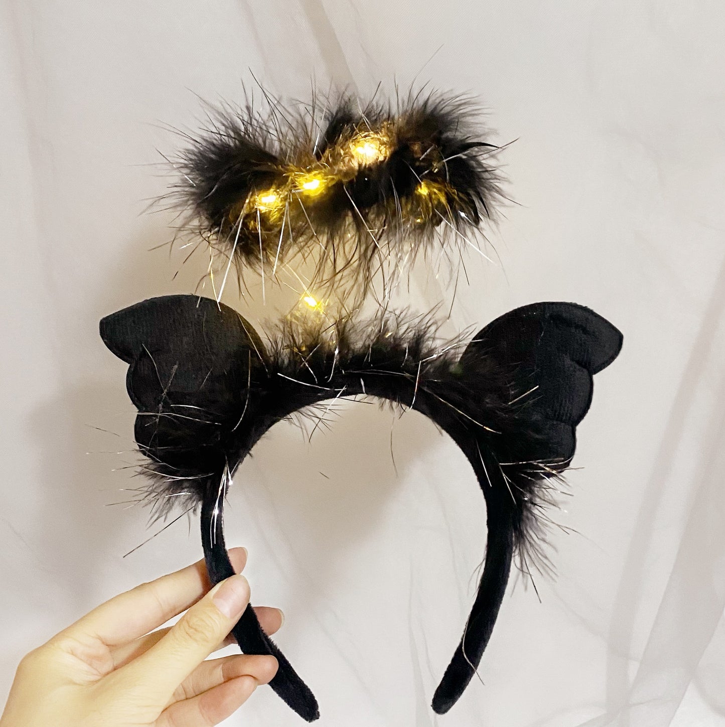 Black angel-themed headband with fluffy cat ears and a small halo adorned with yellow LED lights, held by a hand against a plain white background.