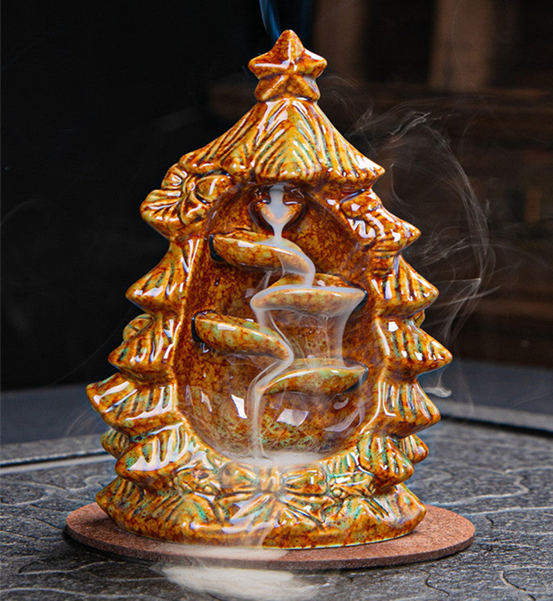 Ceramic incense holder shaped like a Christmas tree with smoke cascading down a spiral path. It's golden-brown in color and set on a dark surface.