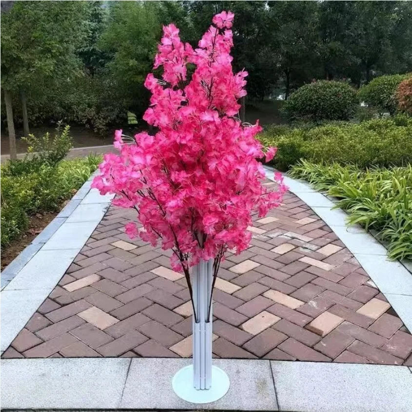 A vibrant bouquet of pink flowers in a white vase stands on a paved garden path, surrounded by lush greenery and trees in the background.