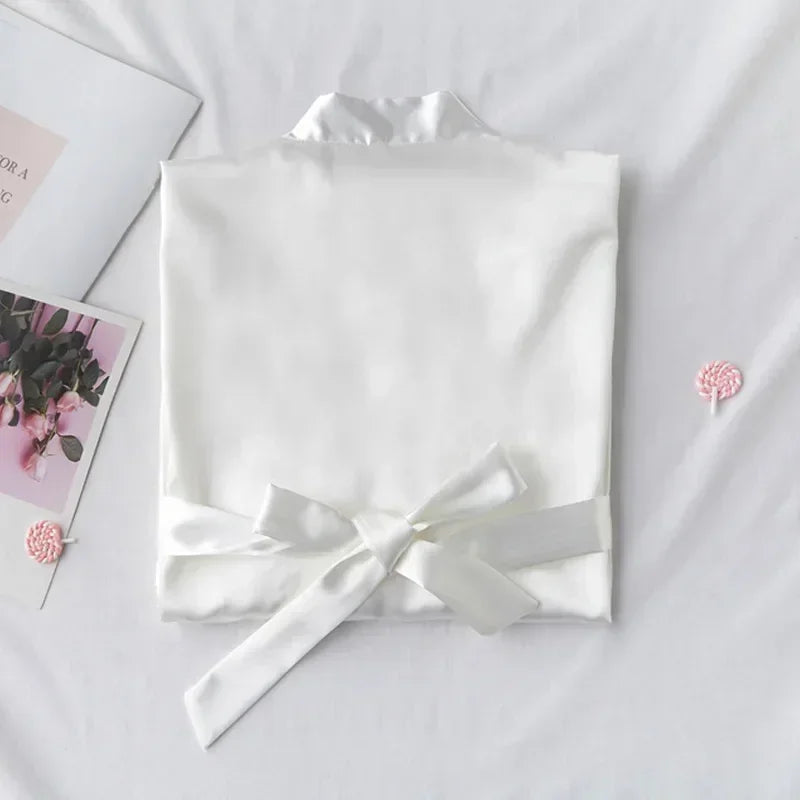 A neatly folded white shirt with a matching satin bow is placed on a white surface, accompanied by a magazine with floral imagery and small pink decorative items.