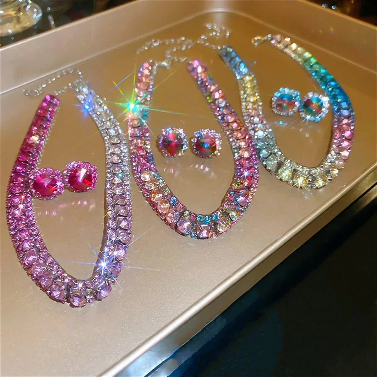 Three sparkling crystal necklaces with matching earrings in pink, multi-color, and rainbow, elegantly displayed on a reflective surface, catching the light beautifully.