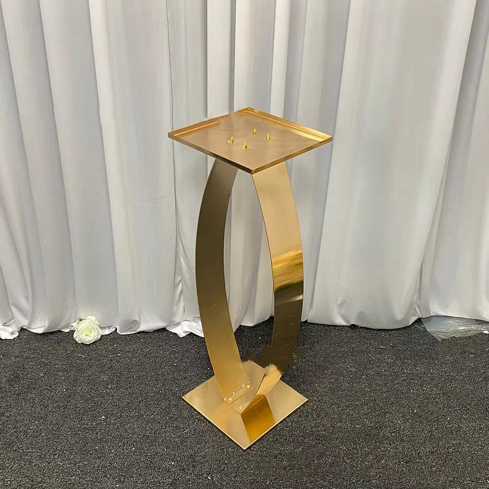 A gold-colored metal pedestal with a curved design stands on a dark carpet, set against white drapes. A single white rose lies on the floor nearby.