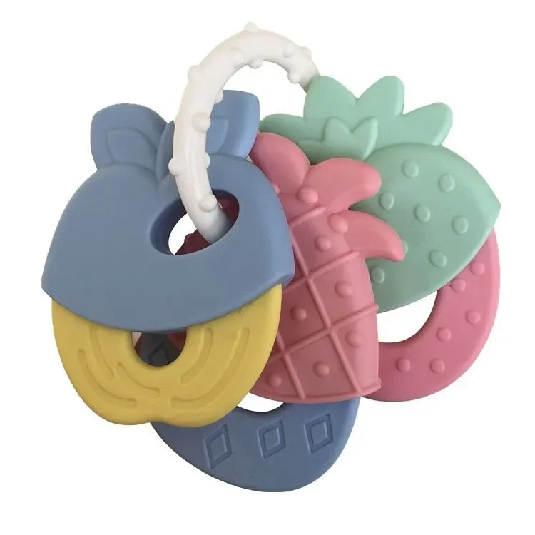 Colorful fruit-shaped baby teething toys on a ring, featuring a blue apple, yellow banana, pink pineapple, and green strawberry. Made of soft, textured silicone.