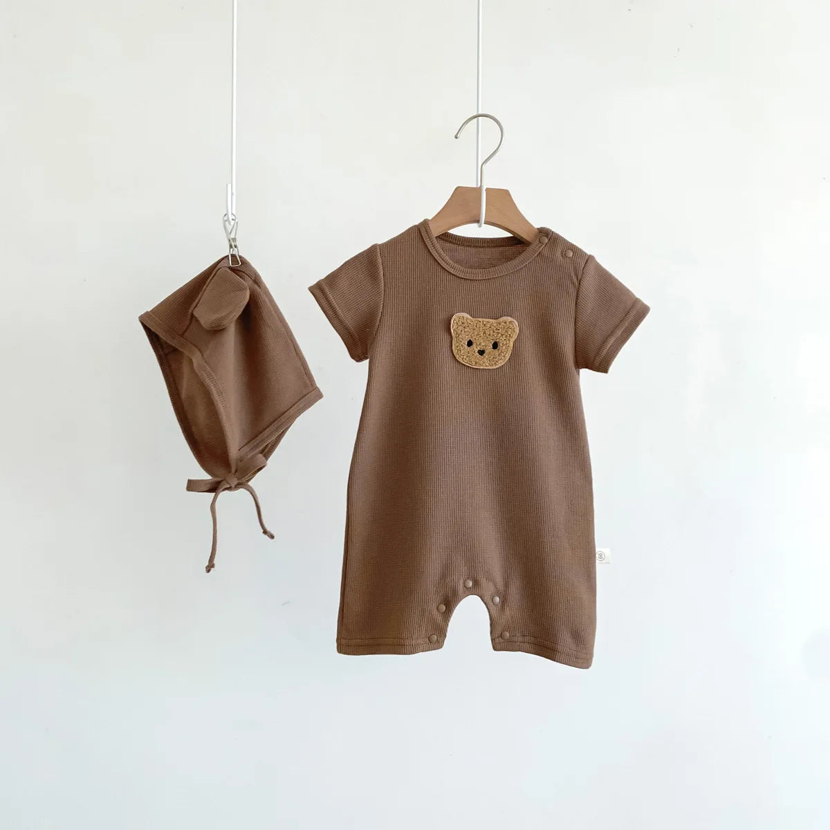 Brown baby romper with short sleeves, featuring a teddy bear design on the chest. A matching brown bonnet with ties hangs nearby, both against a plain white backdrop.