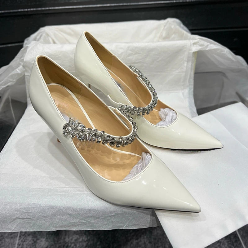 Elegant white pointed-toe heels with crystal embellishments on the strap, resting on a white tissue-lined surface.