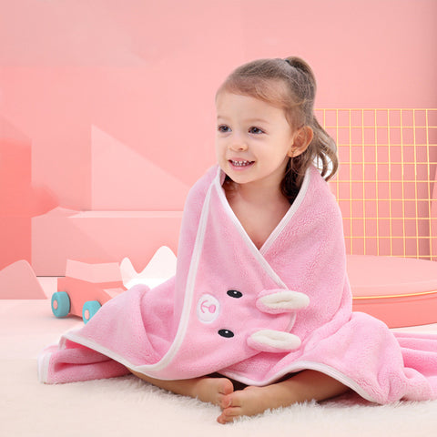 Young child wrapped in a pink animal-themed blanket sits smiling on a soft carpet in a pastel pink room. Toys are scattered in the background, adding a playful touch.