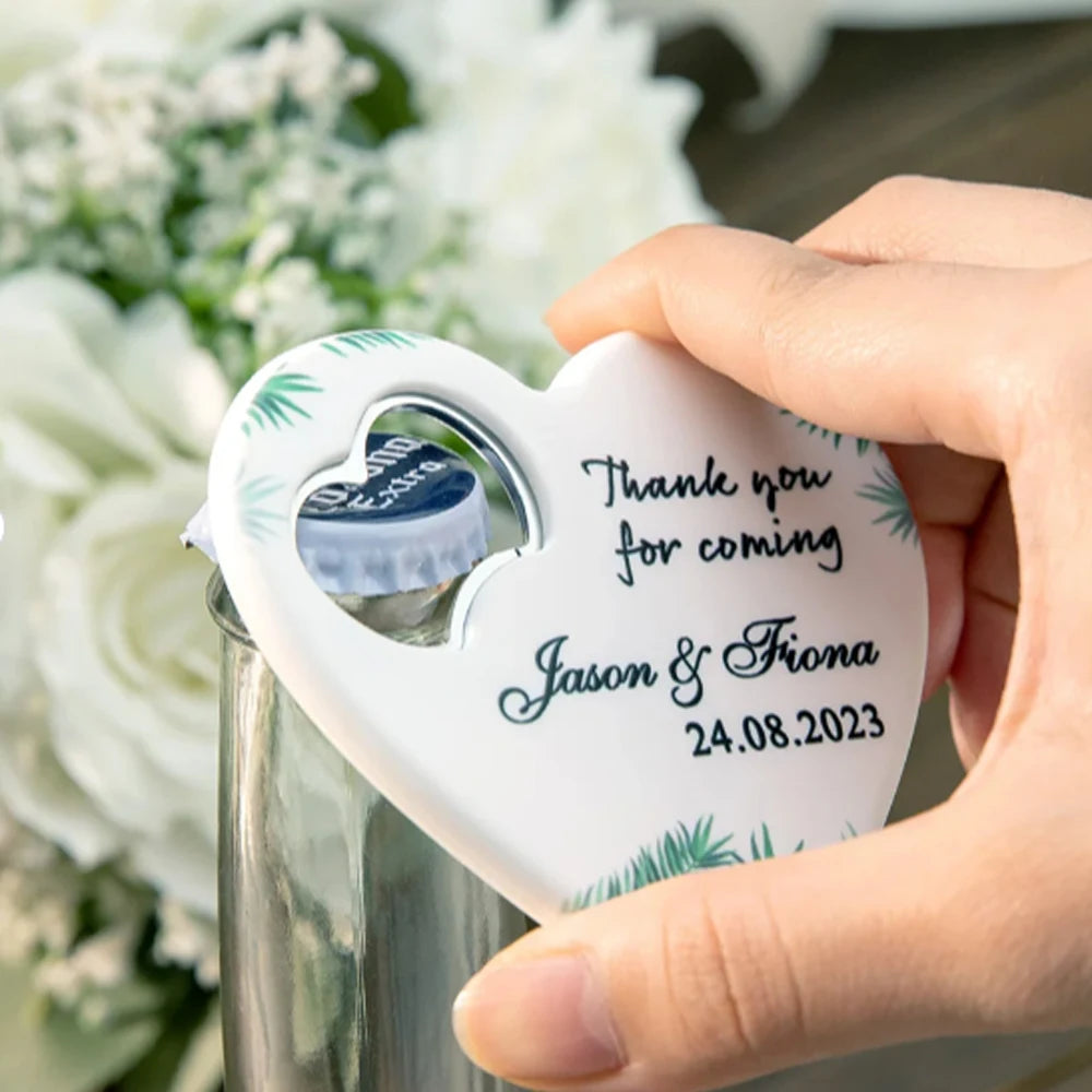 A hand uses a heart-shaped bottle opener with the text "Thank you for coming. Jason & Fiona 24.08.2023" embossed on it. White flowers are blurred in the background.