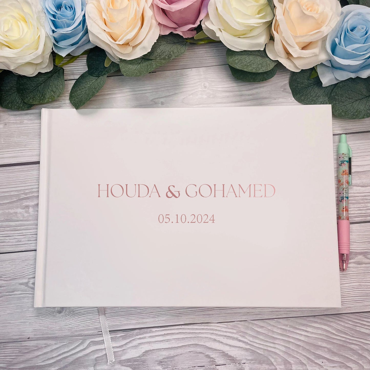 Wedding guest book with "Houda & Gohamed" and date "05.10.2024" on a white cover. Surrounded by pastel roses on wooden surface, a floral pen on the right.