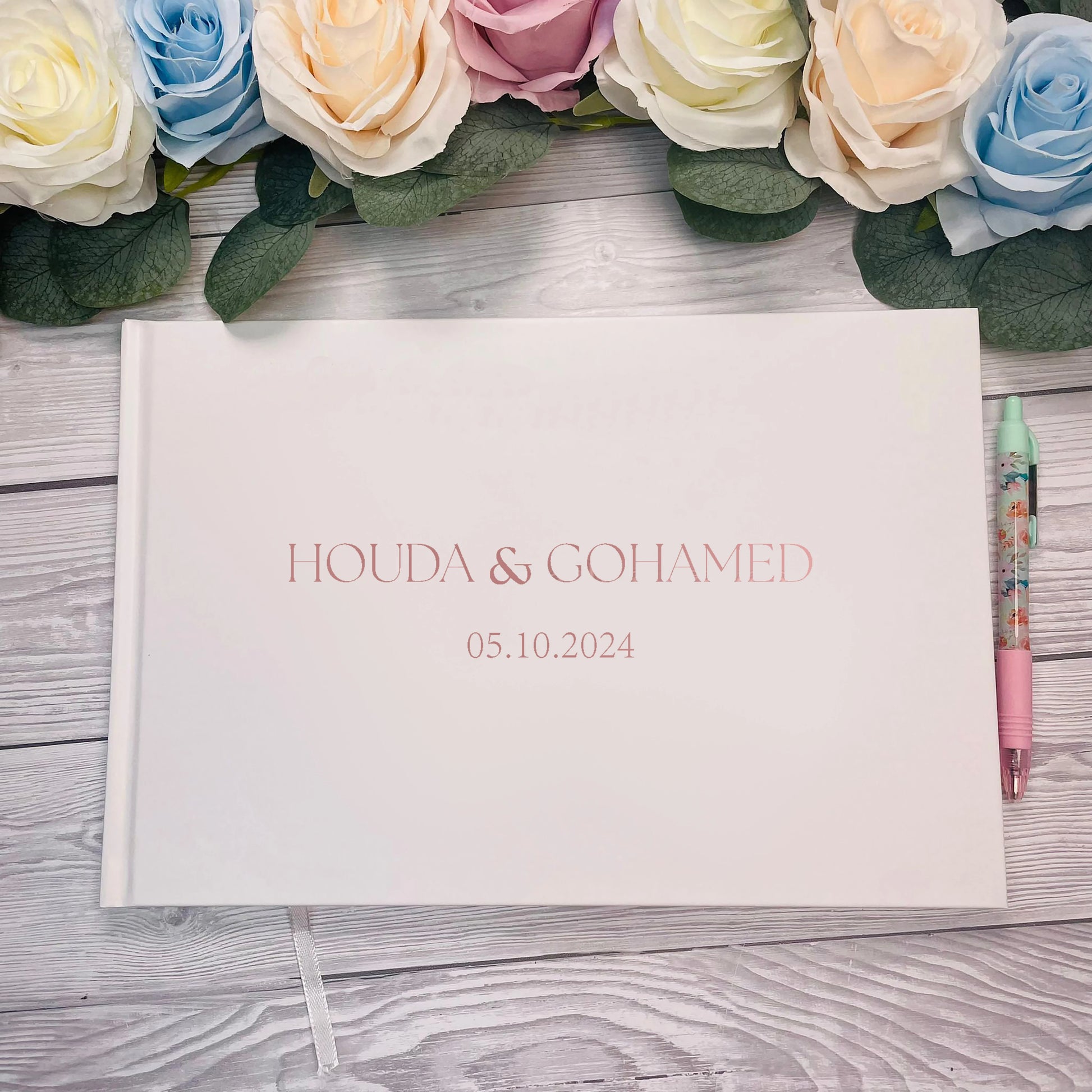 Wedding guest book with "Houda & Gohamed" and date "05.10.2024" on a white cover. Surrounded by pastel roses on wooden surface, a floral pen on the right.