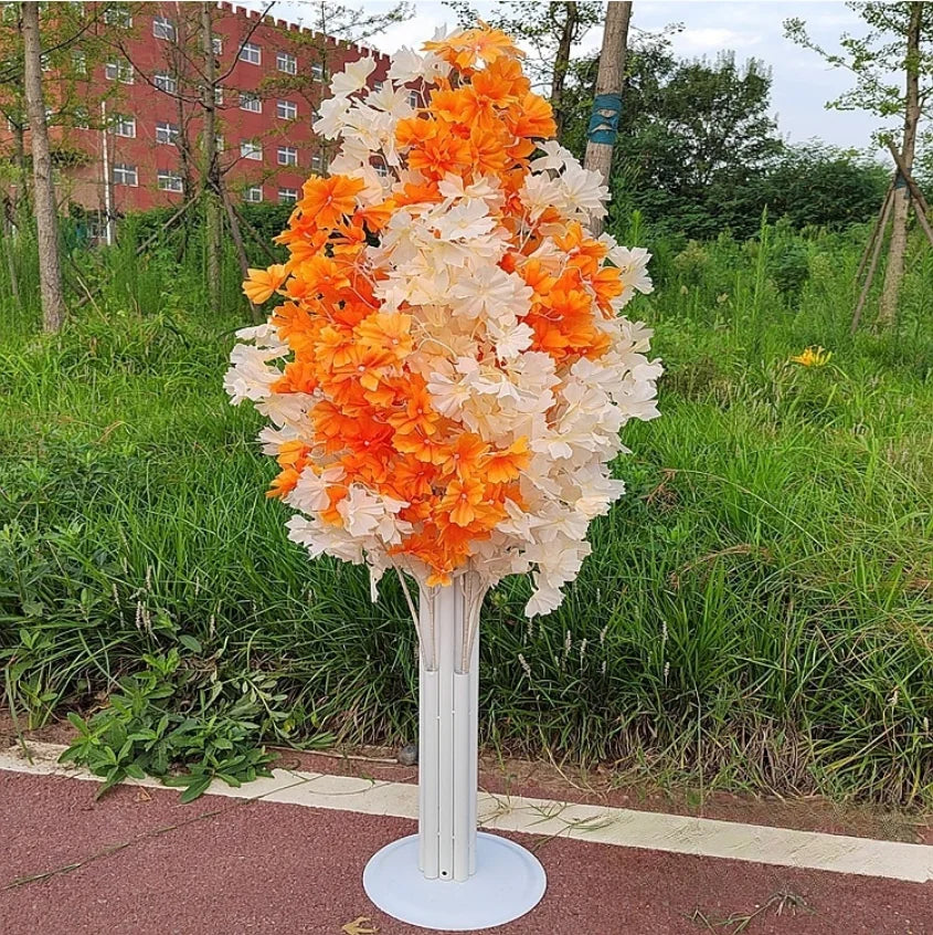 A small artificial tree with orange and white leaves stands on a path, surrounded by green grass. A red brick building is visible in the background.