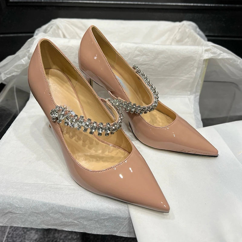 Elegant nude patent leather heels with pointed toes and embellished with a sparkling rhinestone strap, resting on white tissue paper in a black box.