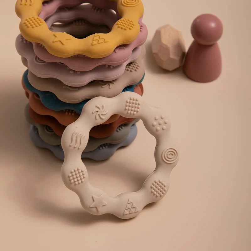 Stack of colorful, textured silicone teething rings in a neat pile with various patterns. Two wooden toys—a faceted block and a pawn-shaped figure—are nearby on a cream surface.