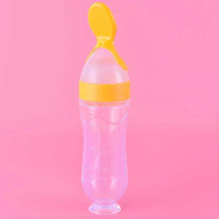Clear baby bottle with yellow spoon top against a pink background. The bottle has measurement markings in milliliters and ounces.