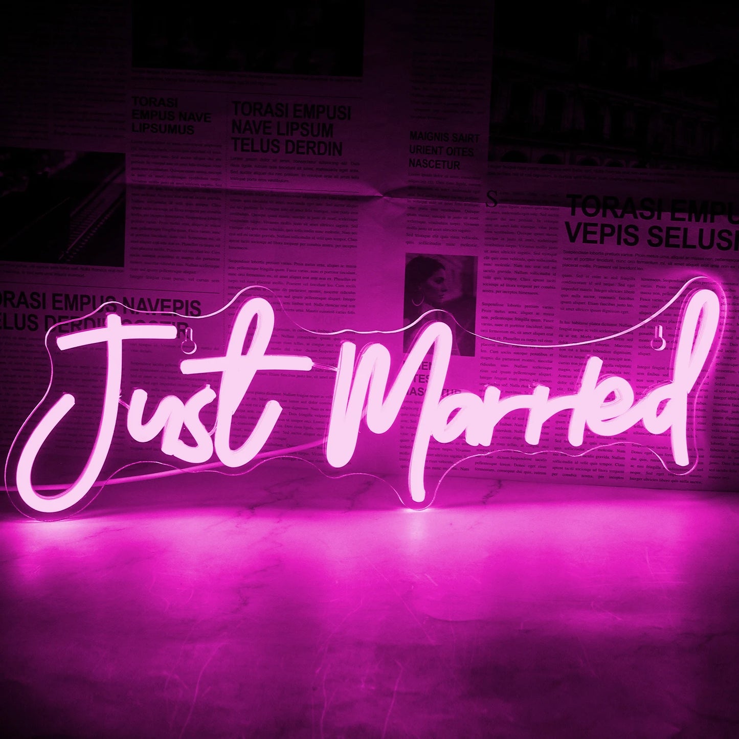 A glowing pink neon sign reads "Just Married" against a backdrop of a newspaper-style wall. The ambient light casts a soft pink hue over the surface below.