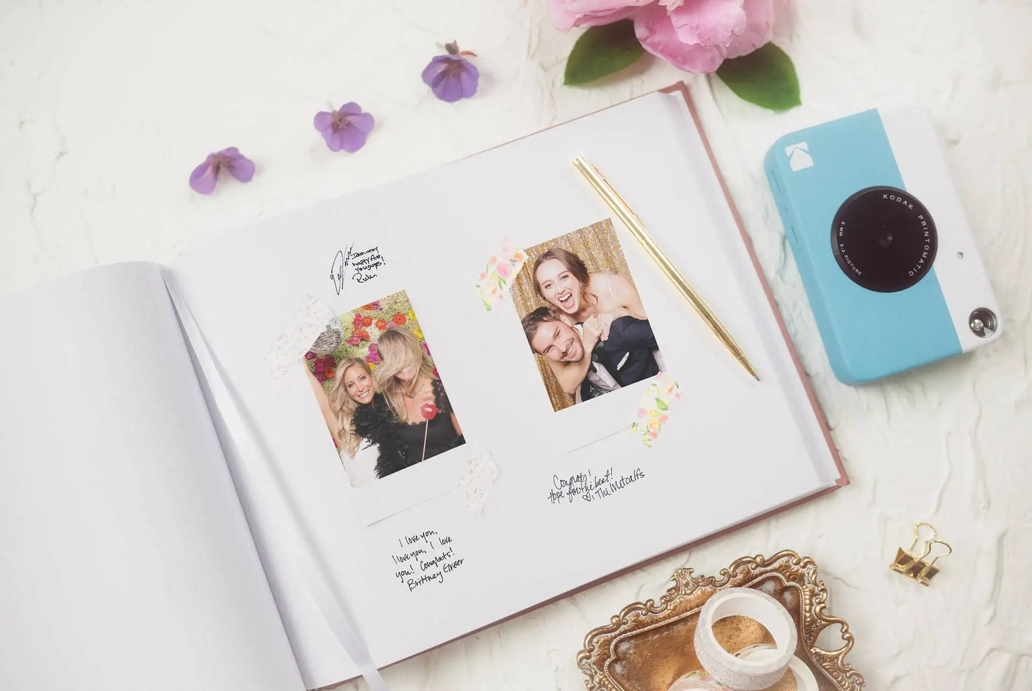 Open scrapbook with two photos taped inside, surrounded by pink flowers and a blue camera. A golden pen rests on the page, next to handwritten messages.