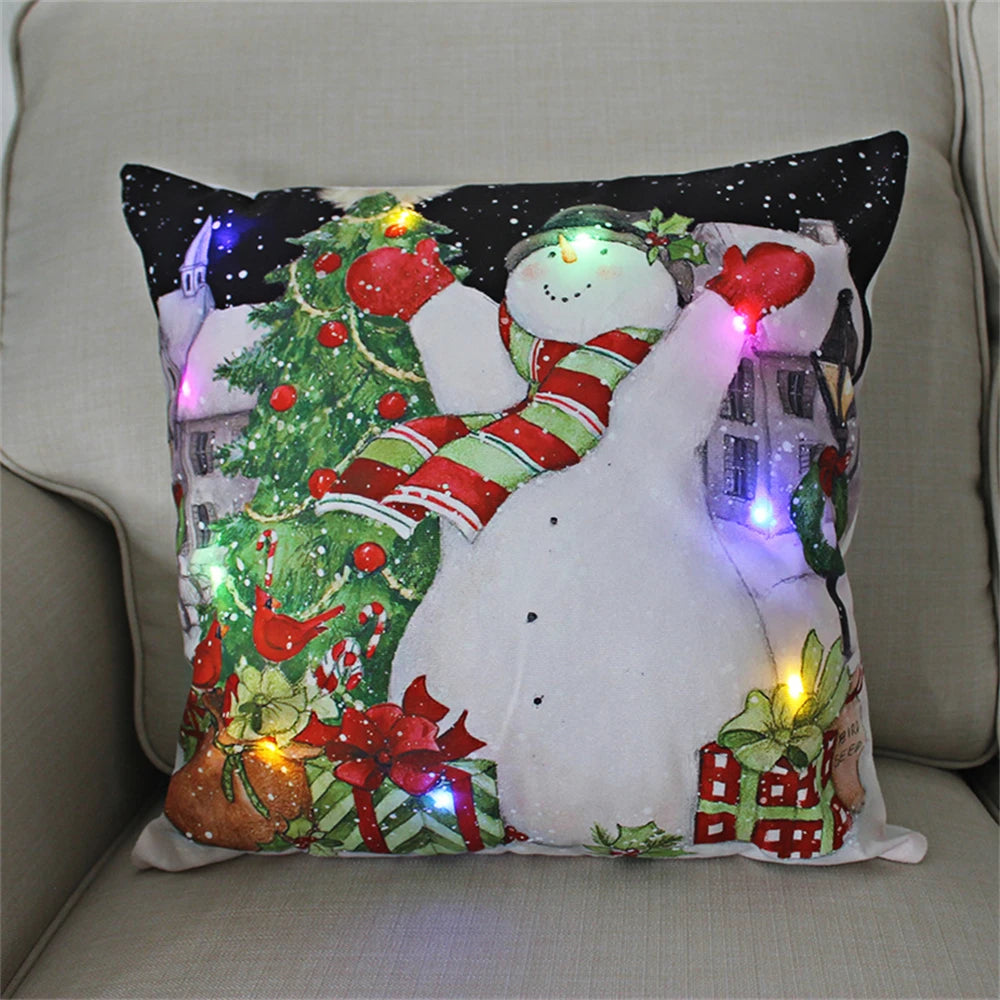 Festive pillow with a snowman, Christmas tree, and presents, adorned with colorful blinking lights. The snowman wears a hat and scarf, sitting on a light-colored couch.