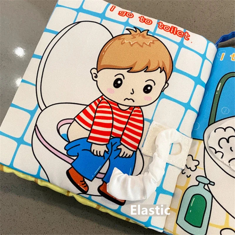 Illustrated fabric page showing a child in a red-striped shirt sitting on a toilet. Text reads, "I go to toilet." An elastic loop is attached to the page.
