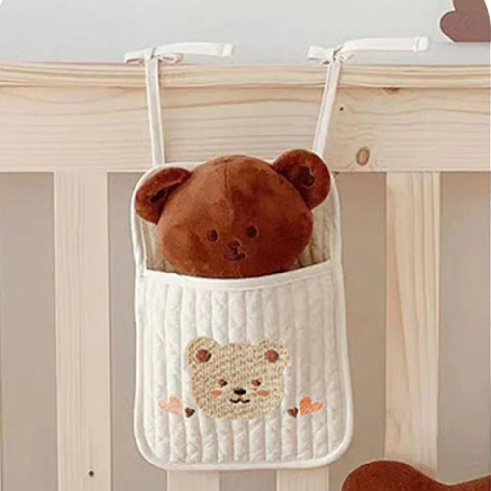 A plush brown bear toy peeks out of a quilted hanging pouch with a teddy bear design, attached to a wooden crib railing.
