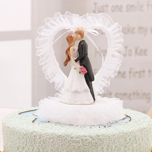 A wedding cake topped with a figurine of a bride and groom kissing, surrounded by a frilly white heart. The bride holds pink roses, and both are in traditional attire.