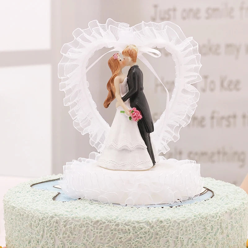 Bride and groom cake topper with a heart-shaped lace background; the bride holds pink flowers, and the groom wears a black suit, both atop a textured white cake.