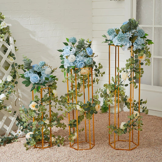 Three gold stands adorned with pastel blue and white flowers and green foliage, set against a white brick wall, creating a fresh and elegant display.