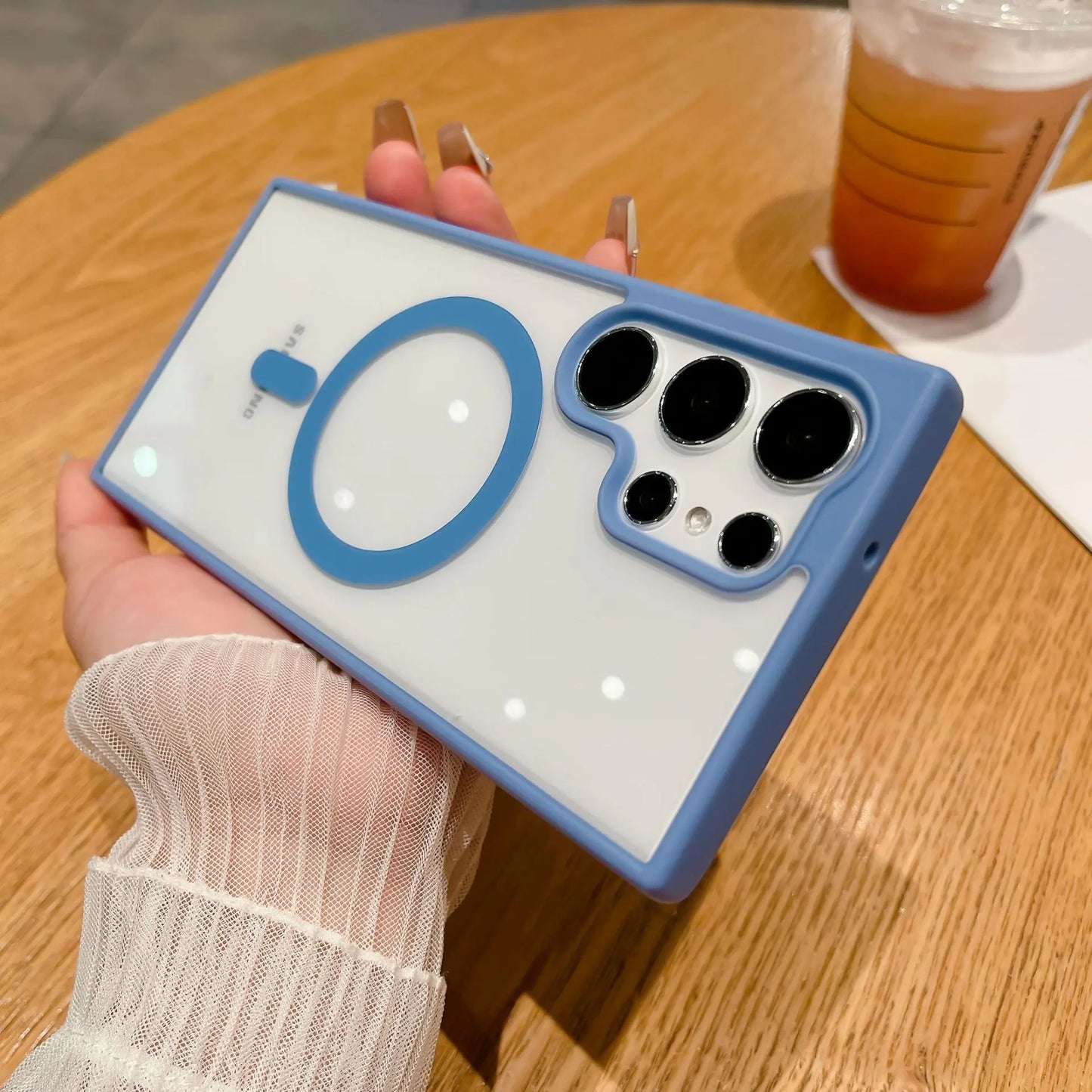 A hand holding a smartphone with a light blue and white case featuring four camera lenses and a circular design on the back. A drink sits on a wooden table in the background.