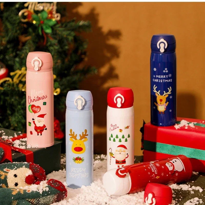 Five festive thermoses with Christmas designs featuring Santa and reindeer are arranged among fake snow and gifts, with a decorated Christmas tree in the background.
