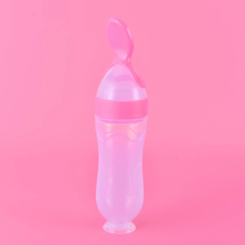 Pink baby feeding bottle with an attached spoon on top, set against a matching pink background. Perfect for easy feeding, blending functionality with cute design.