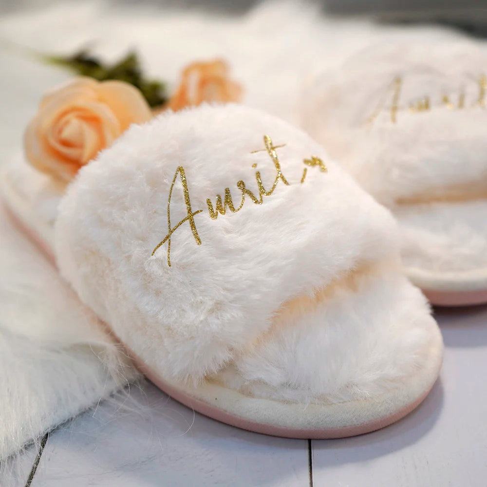 Fluffy white slippers with gold embroidery reading "Amrit." They rest on a soft white surface with peach roses in the background, creating a cozy, elegant setting.