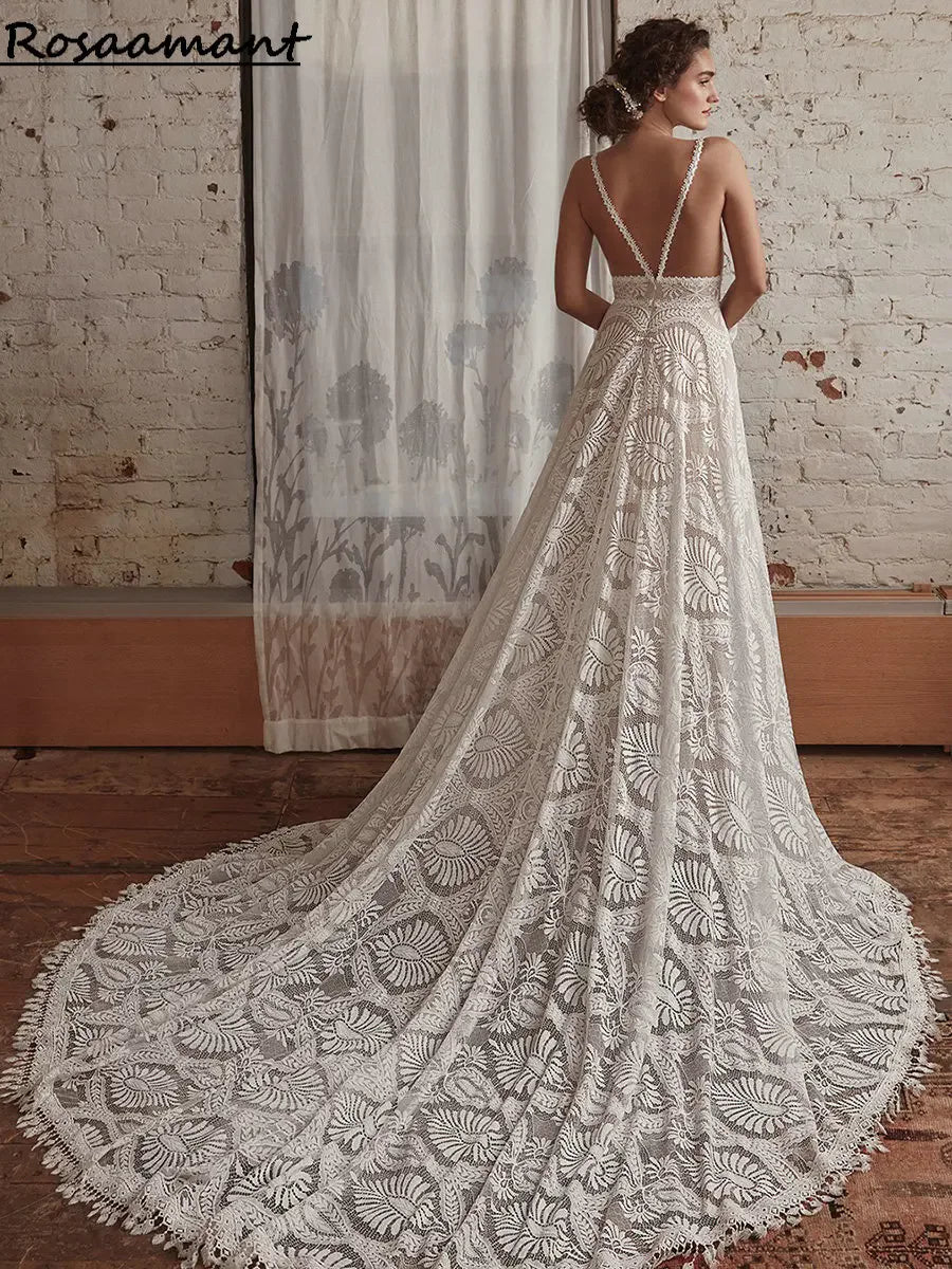 A woman in a lace bridal gown with a long train and floral patterns stands in a rustic room with exposed brick walls and a sheer curtain backdrop.
