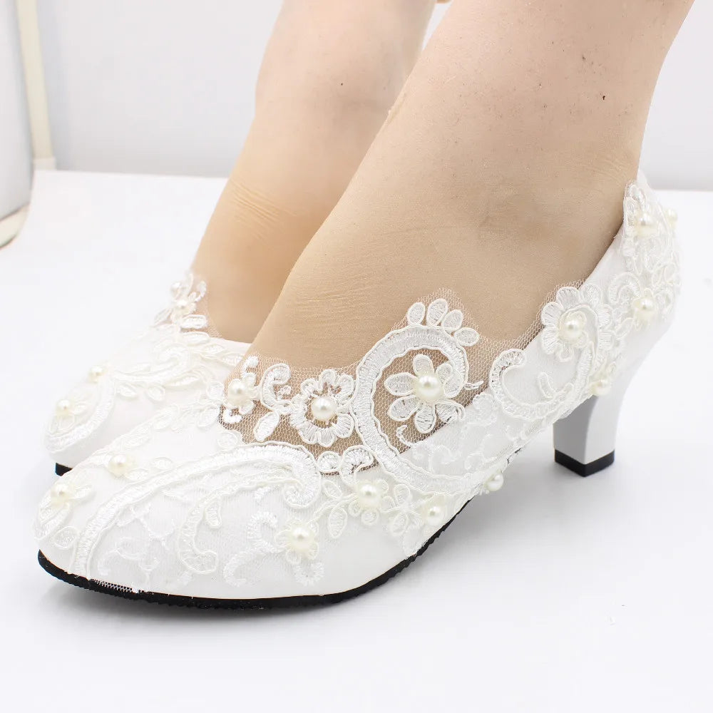 White lace high-heeled shoes adorned with floral embroidery and small pearls, worn by a person with light skin. The shoes have a mesh detail near the ankle and medium heel.