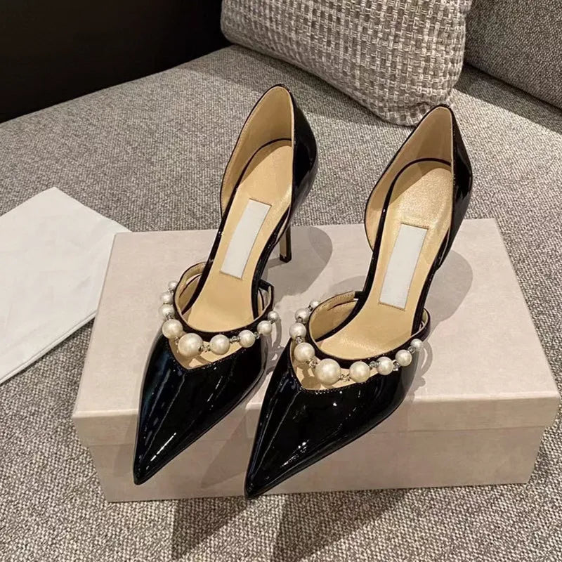 Black patent leather high heels with pointed toes and pearl ankle straps, displayed on a shoebox against a textured sofa backdrop, with a white cloth nearby.