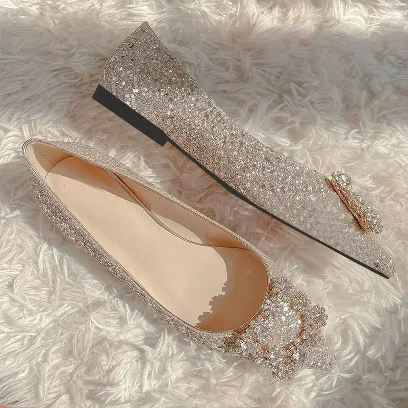 Sparkling silver glitter flats with gem embellishments on the toes, lying on a fluffy white surface. One shoe is upright, the other shows the beige interior.