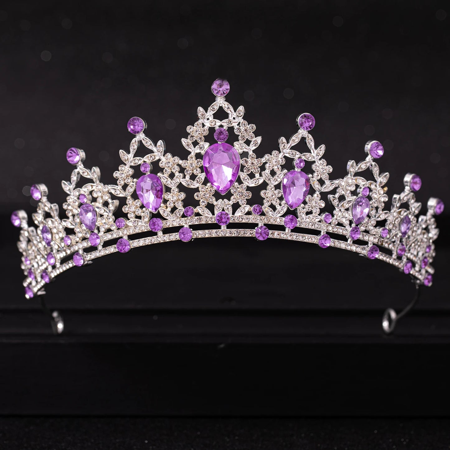 A sparkling tiara adorned with intricate silver details and vibrant purple gemstones, set against a dark background. The design features teardrop and round stones, creating an elegant, regal look.