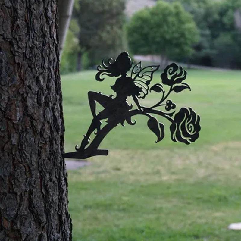Silhouette of a fairy with wings, sitting on a floral design attached to a tree trunk. The background shows a green grassy area with trees.