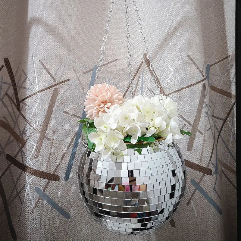 A disco ball used as a hanging flower pot with white and pink flowers, suspended by a chain against a decorative curtain backdrop.