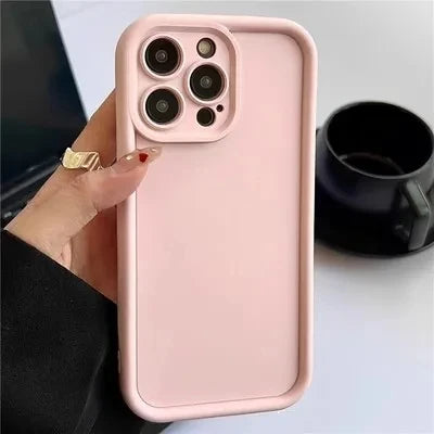 A hand holding a smartphone with a triple-lens camera and a light pink case. A black cup is visible in the background on a white surface.