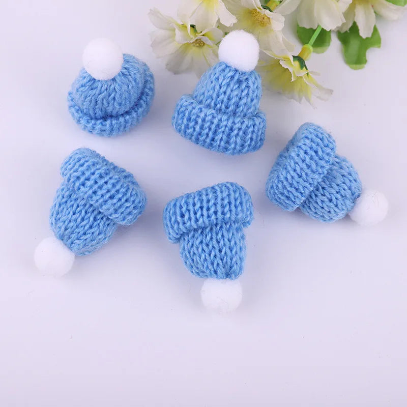 Five small knitted blue hats with white pom-poms are scattered on a white surface, accompanied by white flowers in the background.