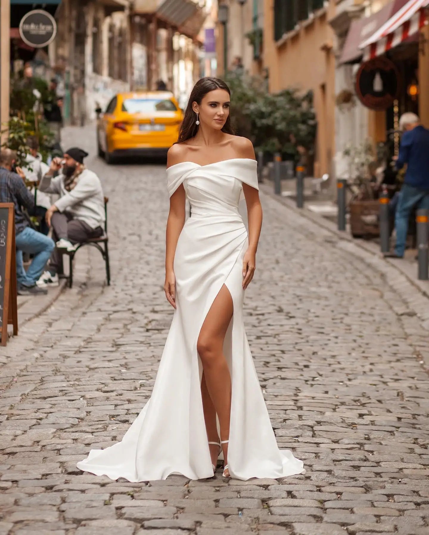 A woman in an elegant white off-shoulder gown with a thigh-high slit walks confidently on a cobblestone street lined with shops and a yellow taxi in the background.