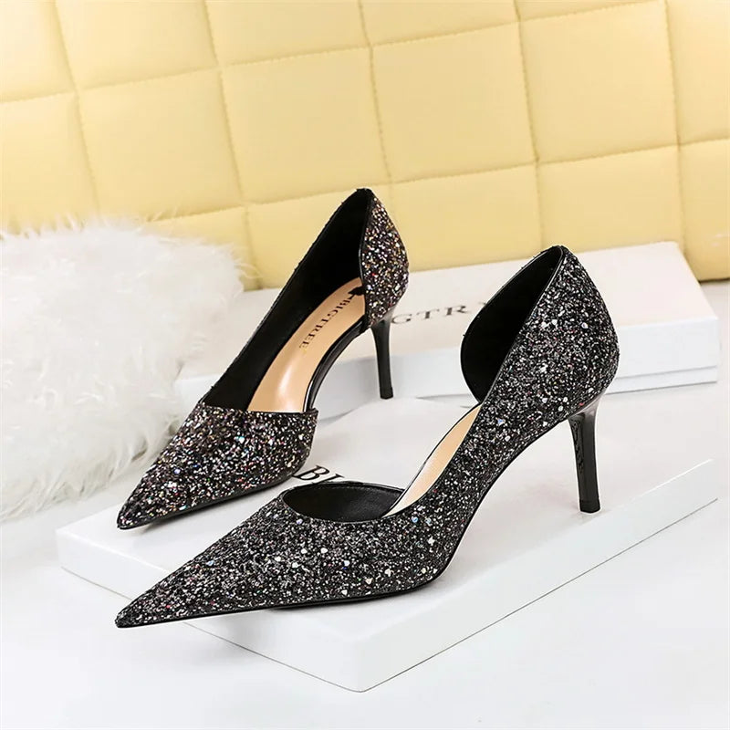 Sparkling black high heels with pointed toes and slim heels on a white box. They have a glittery finish and are displayed against a quilted yellow background.