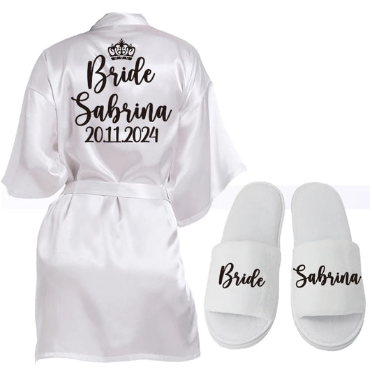 White bridal robe with "Bride Sabrina 20.11.2024" in black script and a small crown graphic. Accompanied by matching white slippers labeled "Bride" and "Sabrina".