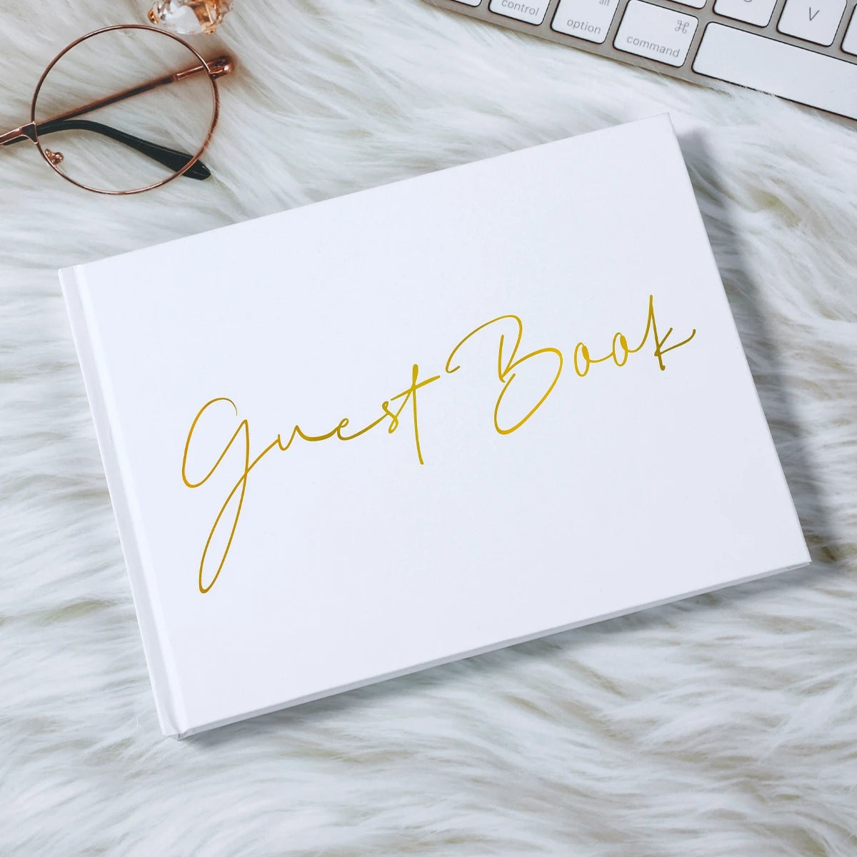 White guest book with "Guest Book" in gold script on a fluffy white surface. Nearby are round glasses and part of a keyboard.