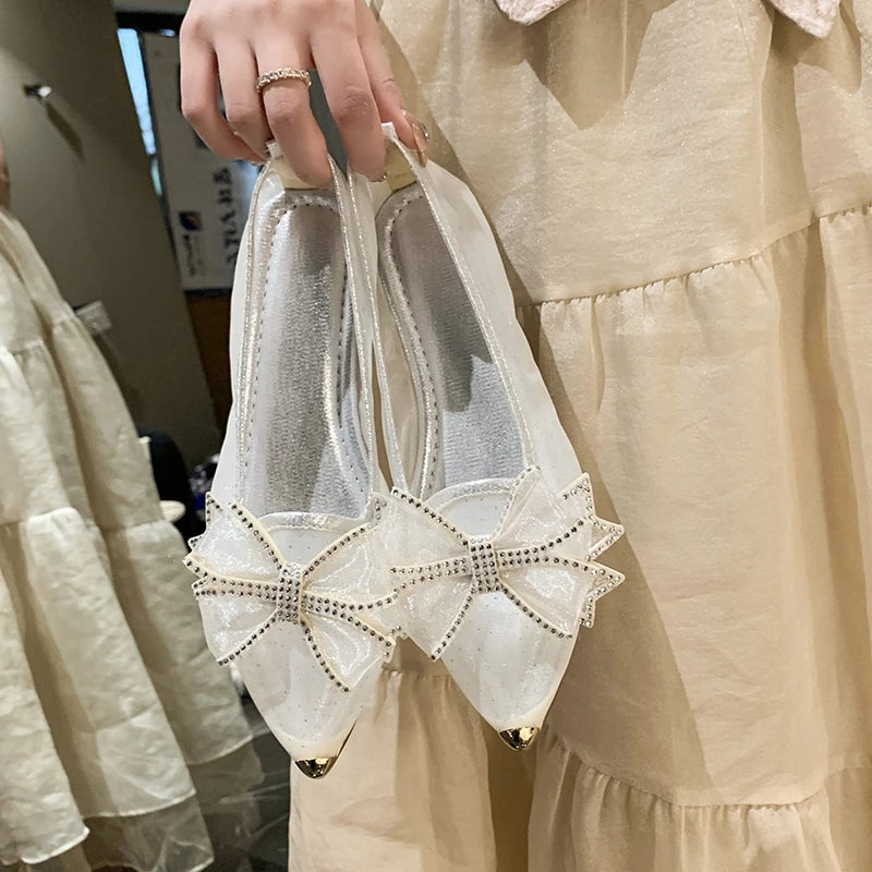 A hand holds a pair of elegant white flats featuring decorative bows with sparkling embellishments and metallic gold-tipped toes, against a beige ruffled dress background.