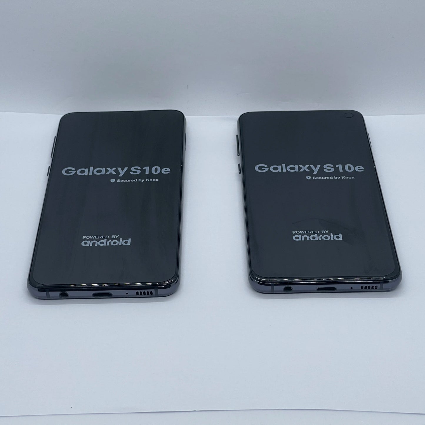 Two Samsung Galaxy S10e smartphones are lying side by side face-up on a white surface, displaying the startup screen with "Secured by Knox" and "Powered by Android" logos.