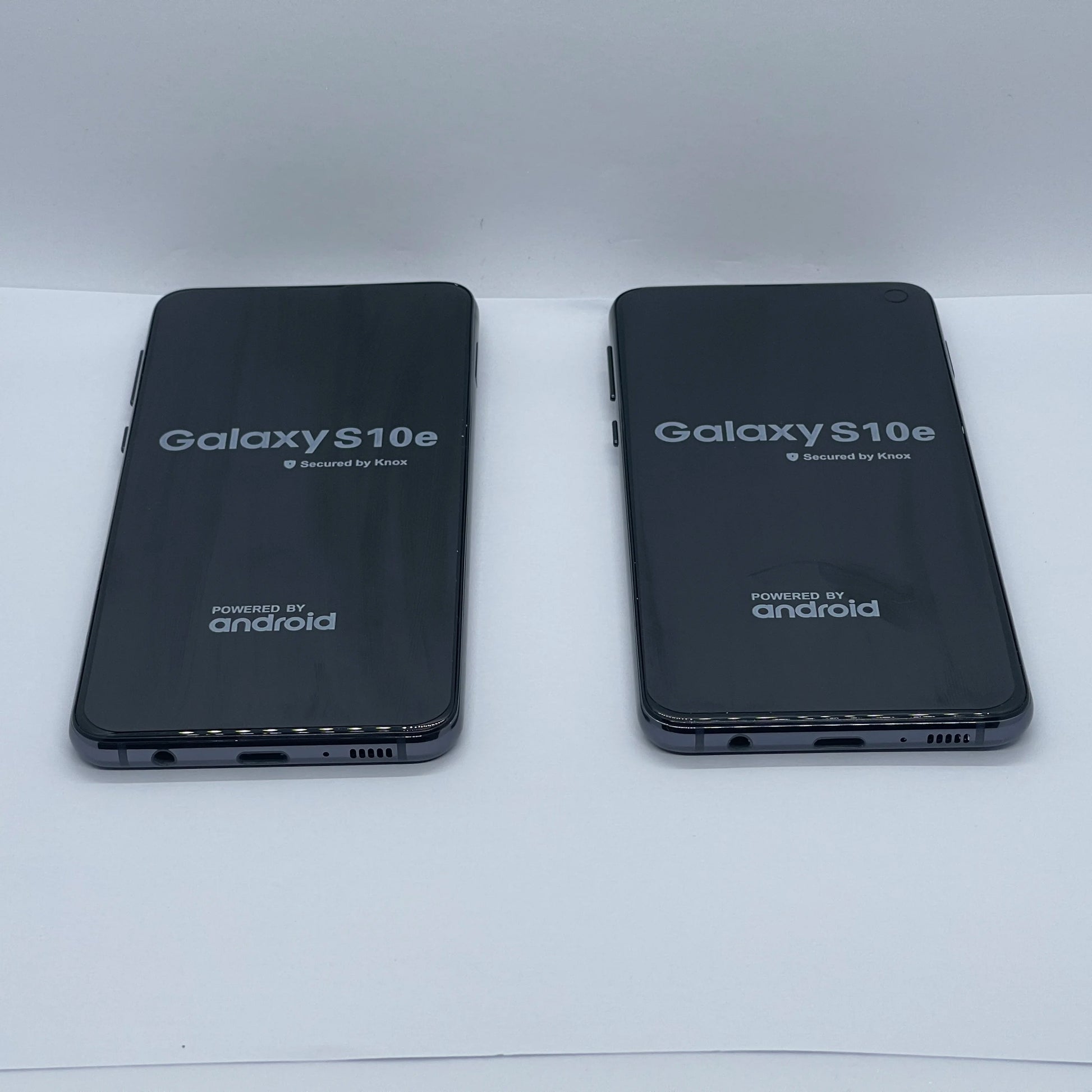 Two Samsung Galaxy S10e smartphones are lying side by side face-up on a white surface, displaying the startup screen with "Secured by Knox" and "Powered by Android" logos.