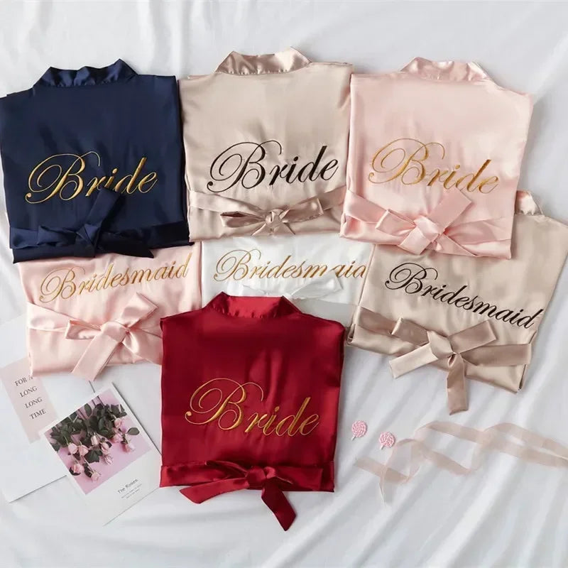 Silk robes displayed on a white surface, labeled "Bride" and "Bridesmaid" in various colors: navy, blush, red, and champagne. A card and pink flowers are placed nearby.