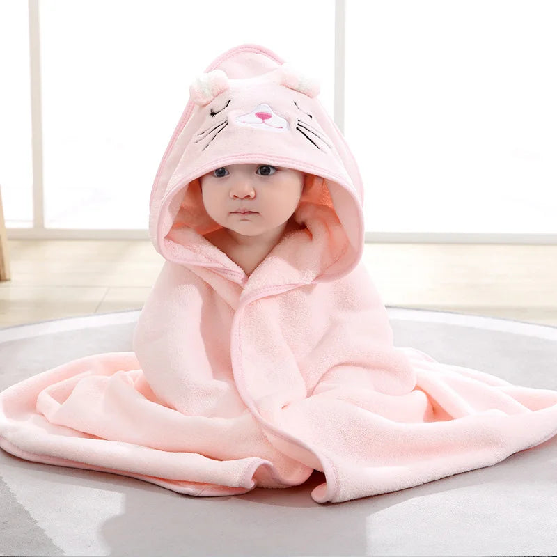 A baby wrapped in a soft pink blanket with a hood featuring cat ears and a cute face, sitting on a light-colored floor.