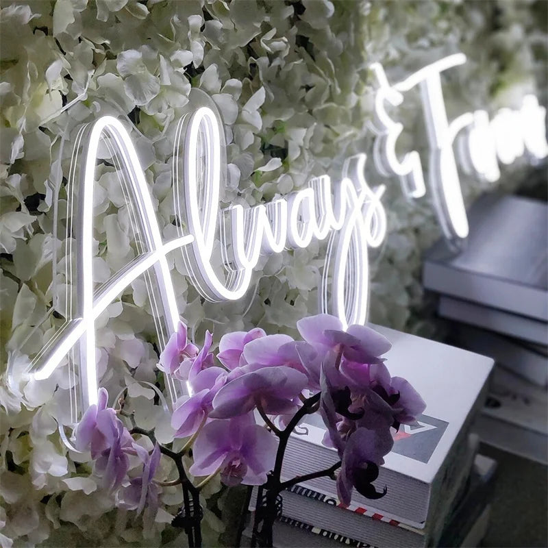Neon sign reading "Always and Forever" on a backdrop of white flowers and purple orchids. Stacked books are visible in the lower right corner.